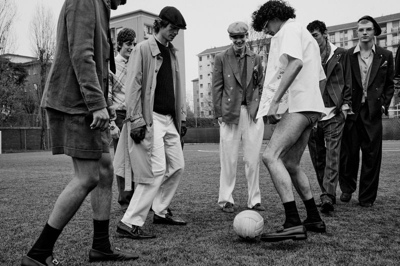 Group of male models in vintage attire gathered around a soccer ball on a grassy field with apartment buildings in the background.