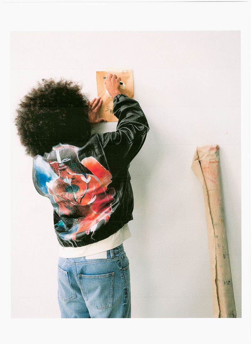 Rear view of a person with an afro hairstyle wearing a painted black jacket and blue jeans drawing on a white wall