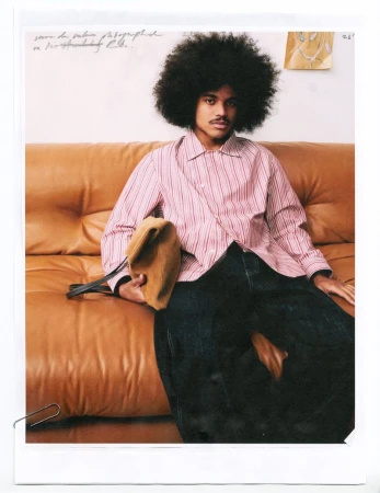Portrait of Samuel de Saboia with a large afro seated on a tan leather sofa wearing a striped shirt and dark trousers.