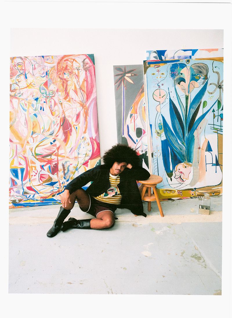 Portrait of a man with an afro hairstyle seated on the floor surrounded by large colorful paintings in an artist studio