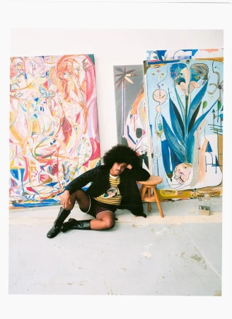 Portrait of a man with an afro hairstyle seated on the floor surrounded by large colorful paintings in an artist studio