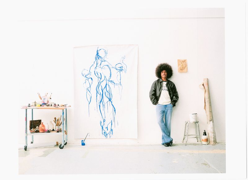 Man with afro hair wearing a dark leather jacket and denim jeans standing in a white studio next to large blue artwork.