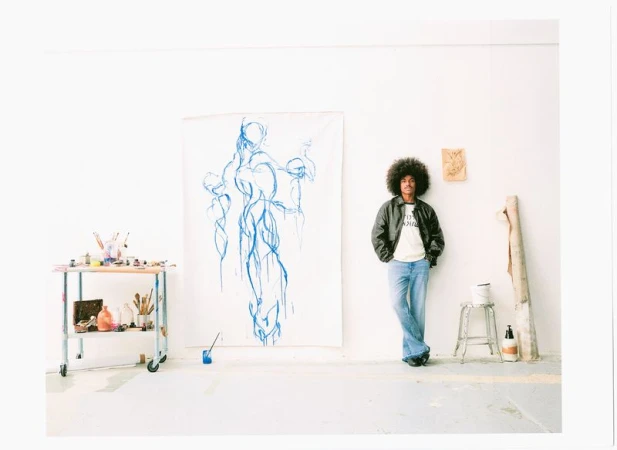 Man with afro hair wearing a dark leather jacket and denim jeans standing in a white studio next to large blue artwork.