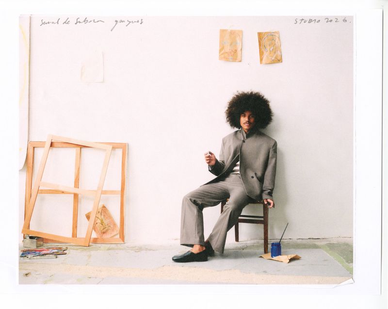 Man with large afro wearing a grey suit jacket and matching trousers seated on a chair in a sparse studio space.