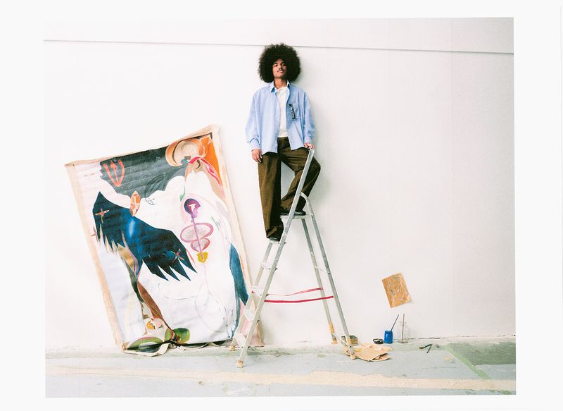 A Black man with an afro standing on a stepladder leaning against a plain wall next to a large abstract painting.