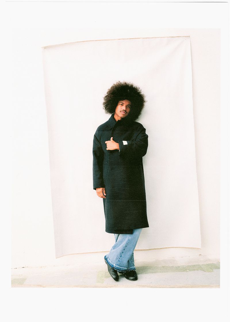 Man with afro posing in a long black coat and blue jeans against a white backdrop, looking at the camera with neutral expression.