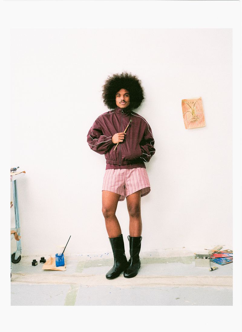 Man with afro hair wearing a burgundy track jacket and striped shorts standing against a white wall