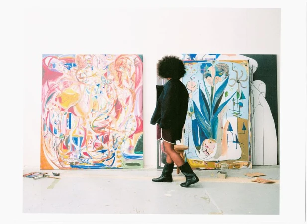 Person with an afro hairstyle wearing a dark jacket and black boots walking past large colorful paintings