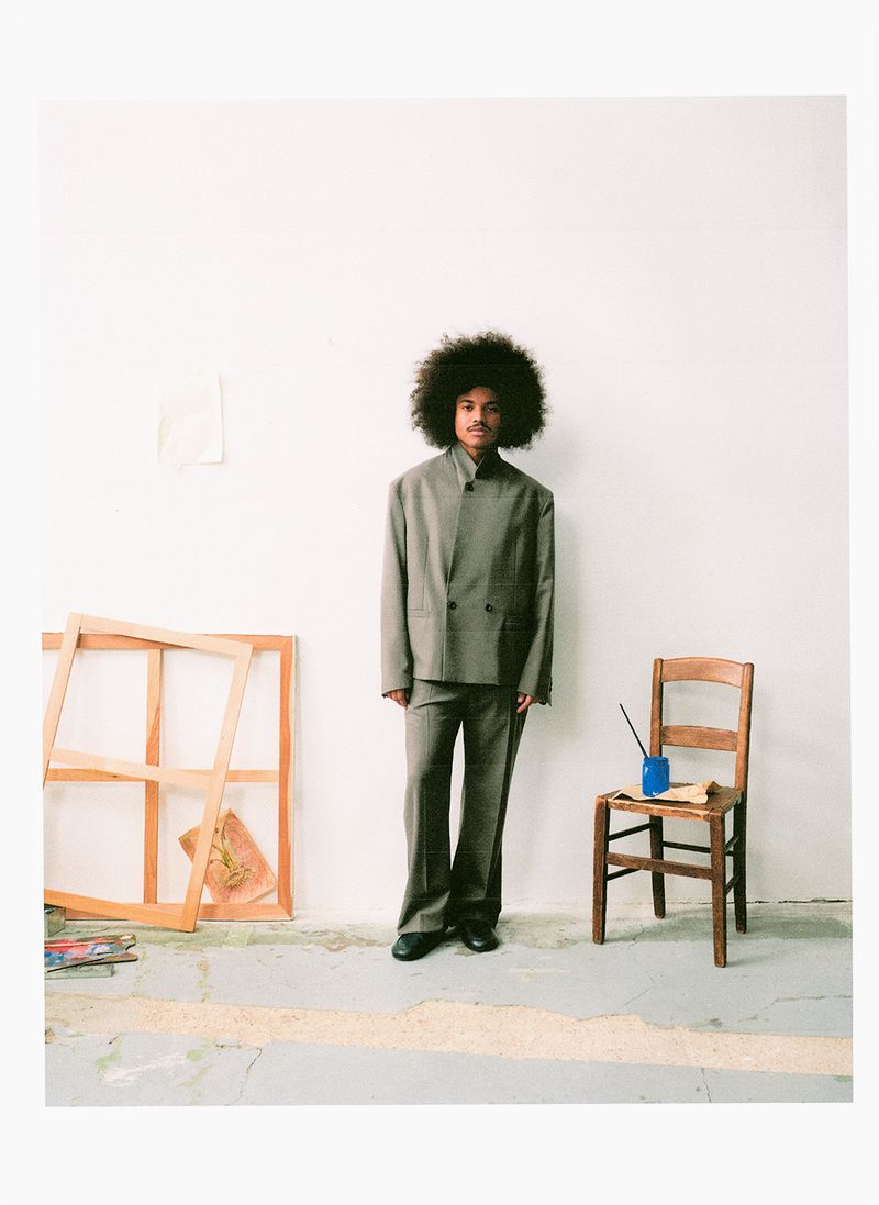 Full-length portrait of a man in a grey suit with an afro hairstyle standing in an artist studio with a wooden chair and empty frames.
