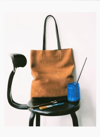 Brown leather tote bag resting on a black chair with paintbrushes and a blue jar against a white wall.