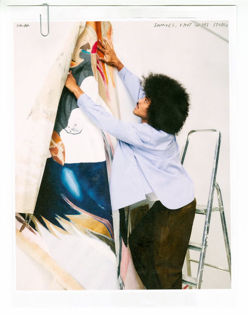 Man with afro hair wearing a light blue shirt hanging a large colorful painted canvas in a studio.