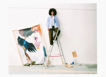 A Black man with an afro standing on a stepladder leaning against a plain wall next to a large abstract painting.