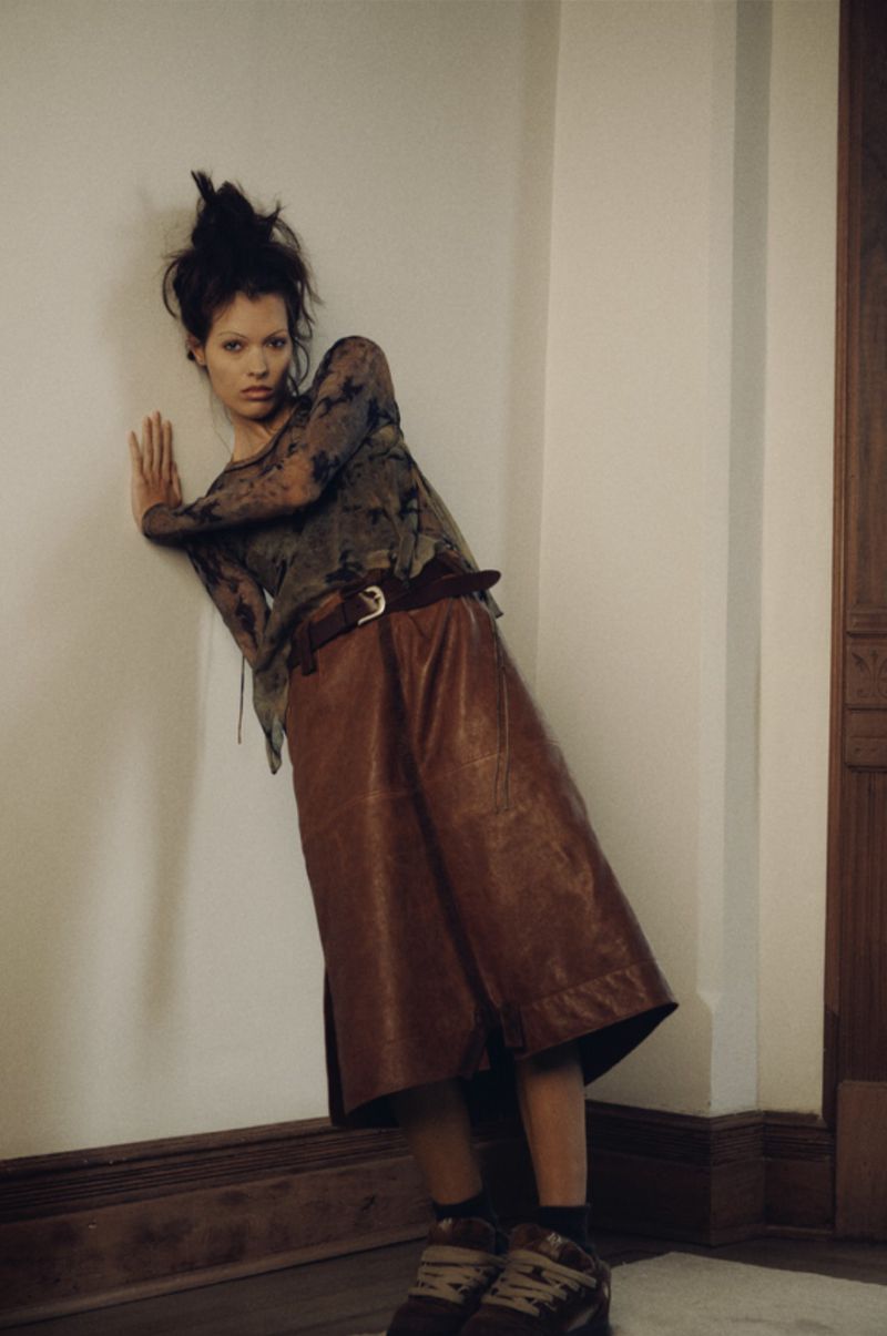 A young woman with dark hair poses in front of a white wall, wearing a long-sleeved top, a leather skirt with a belt, and chunky sneakers.