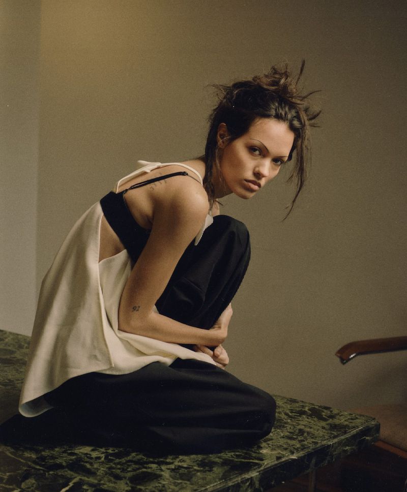 Model crouched on a green marble table wearing a sleeveless white top and black trousers against a neutral wall