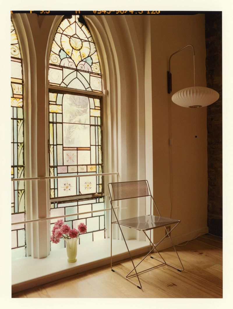 Wire frame modern chair near tall stained glass windows with a vase of pink flowers on the sill.