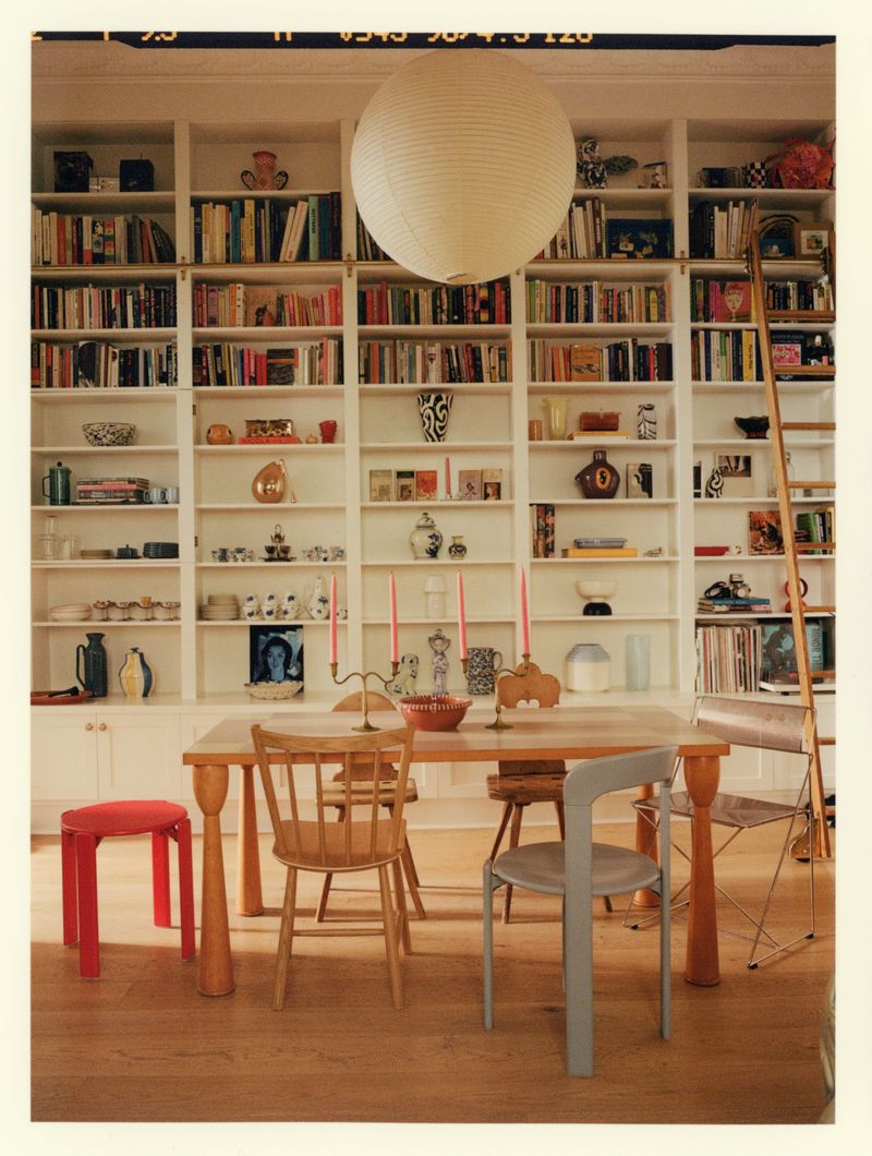 Interior dining space featuring a wooden table surrounded by mismatched chairs, large floor-to-ceiling white bookshelves, and a spherical pendant lamp.