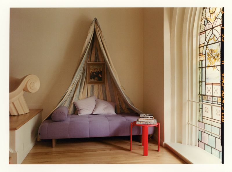 Purple daybed with pillows beneath a draped striped canopy next to a stained glass window.