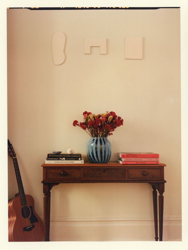 Wooden console table holding a blue striped vase with red flowers, stacked books, and an acoustic guitar leaning on the wall.