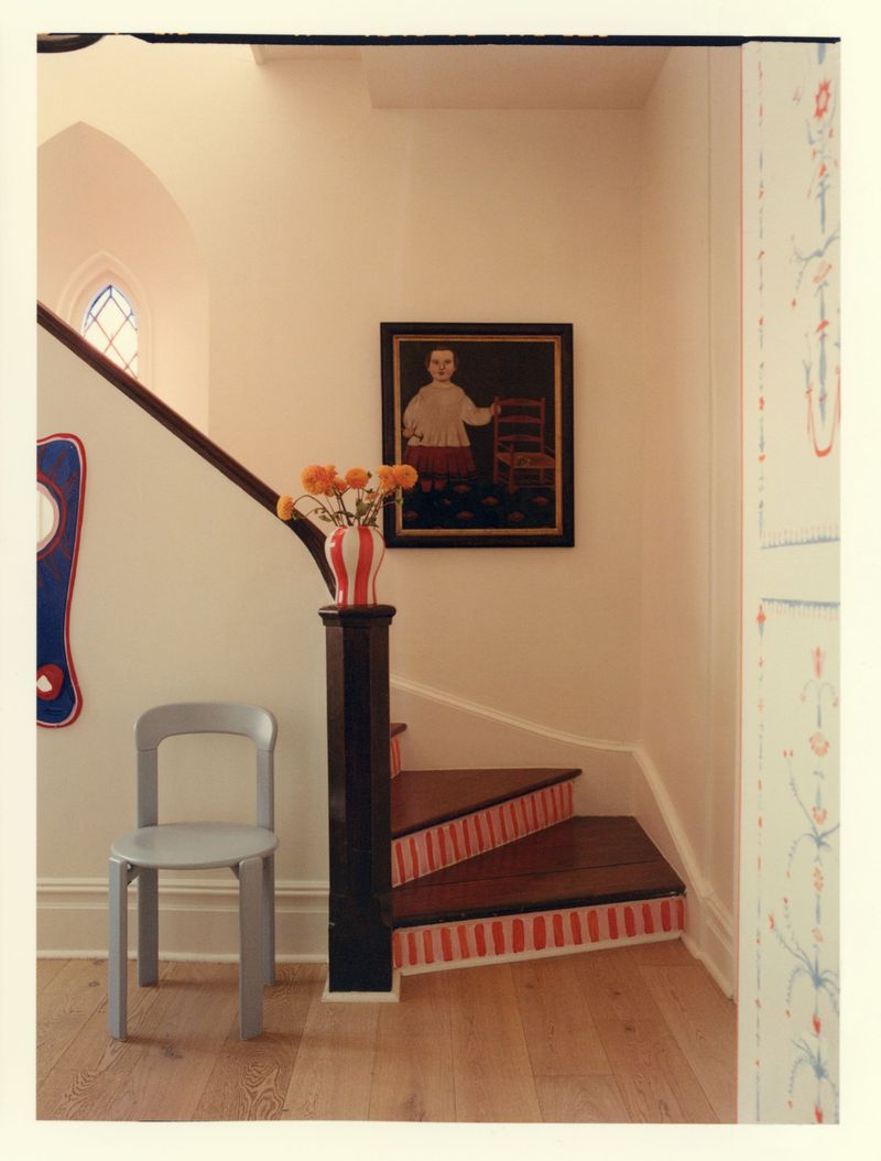 Interior shot featuring a light grey wooden chair beside a staircase with striped risers and a framed portrait on the wall.