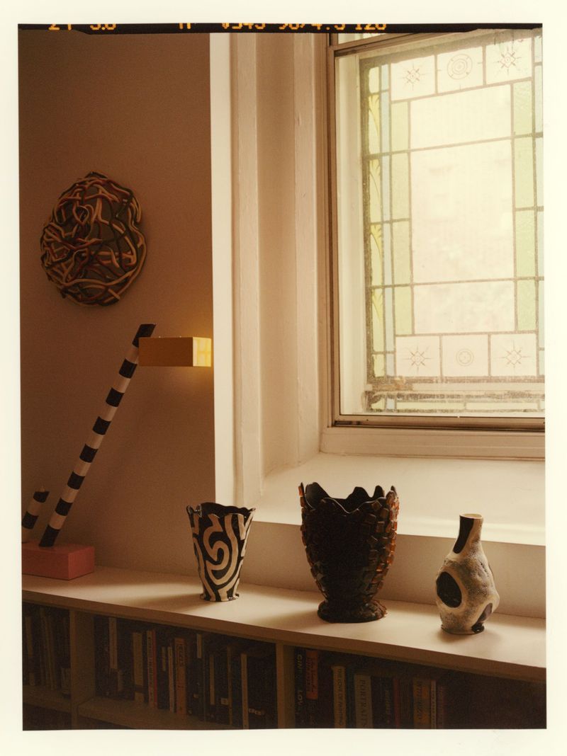 Still life featuring art objects on a shelf in front of a window and bookshelves: vases, sculpture, and painted decoration.
