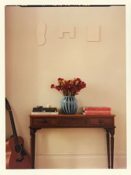 Wooden console table holding a blue striped vase with red flowers, stacked books, and an acoustic guitar leaning on the wall.