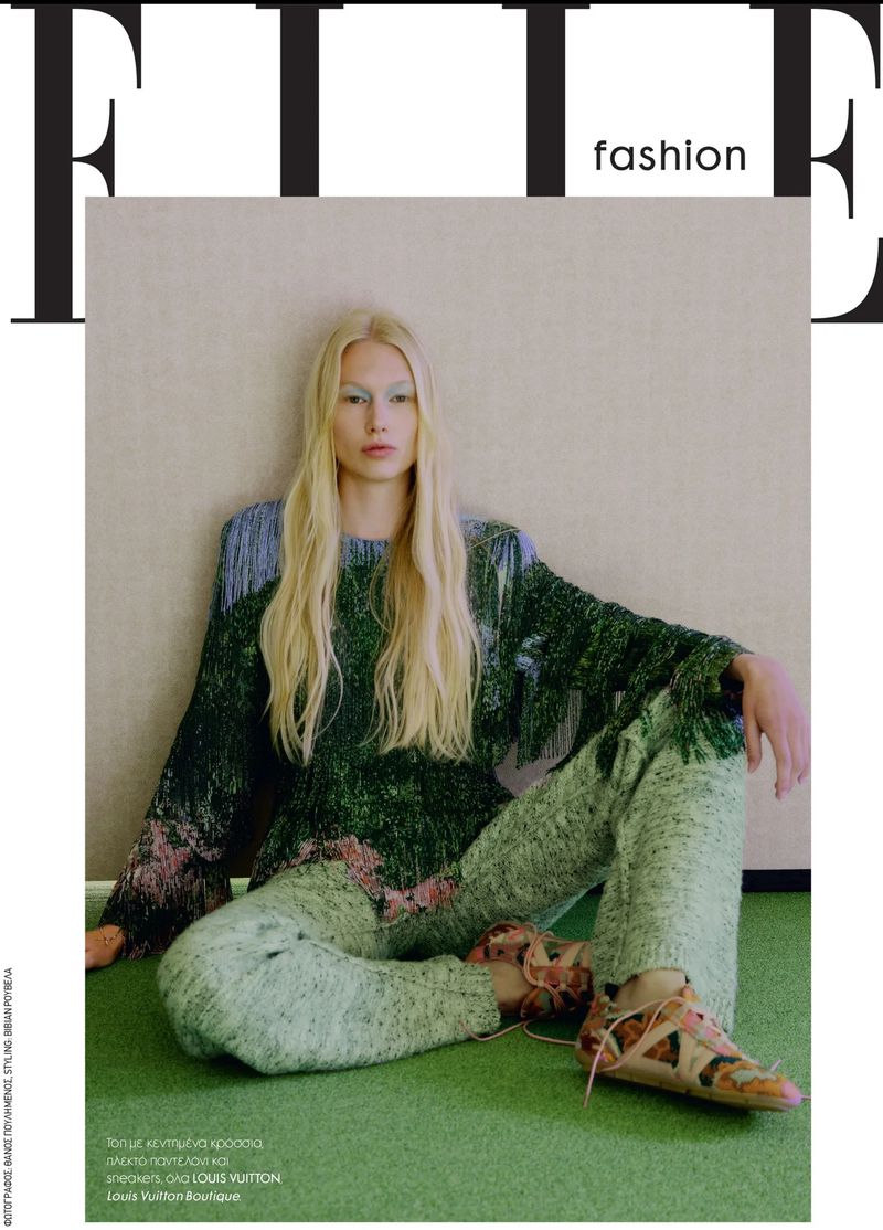 Blonde female model wearing green knit sweater and patterned trousers seated against a textured wall for Elle Greece Quiet Bloom editorial