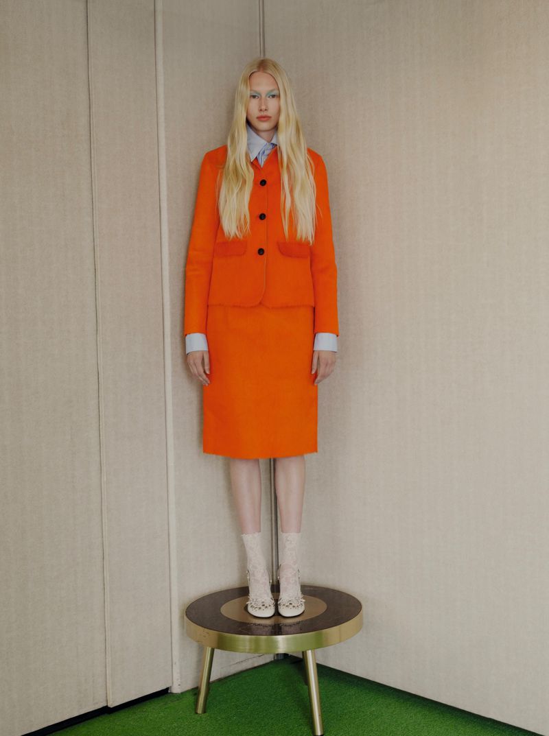 Blonde model in a vibrant orange skirt suit standing on a small round pedestal in an interior corner setting.