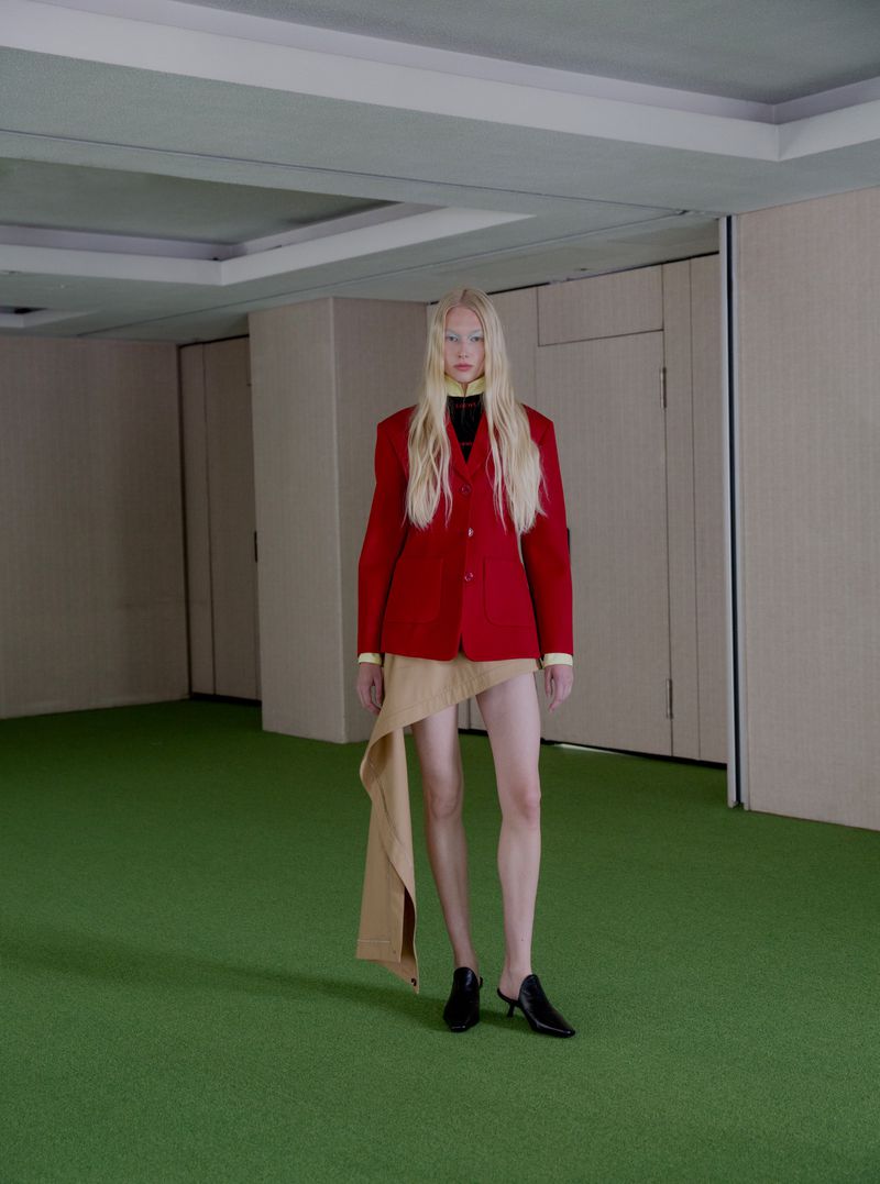 Model with long blonde hair wearing a red blazer and beige skirt standing in a room with green floor