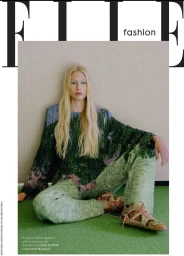 Blonde female model wearing green knit sweater and patterned trousers seated against a textured wall for Elle Greece Quiet Bloom editorial