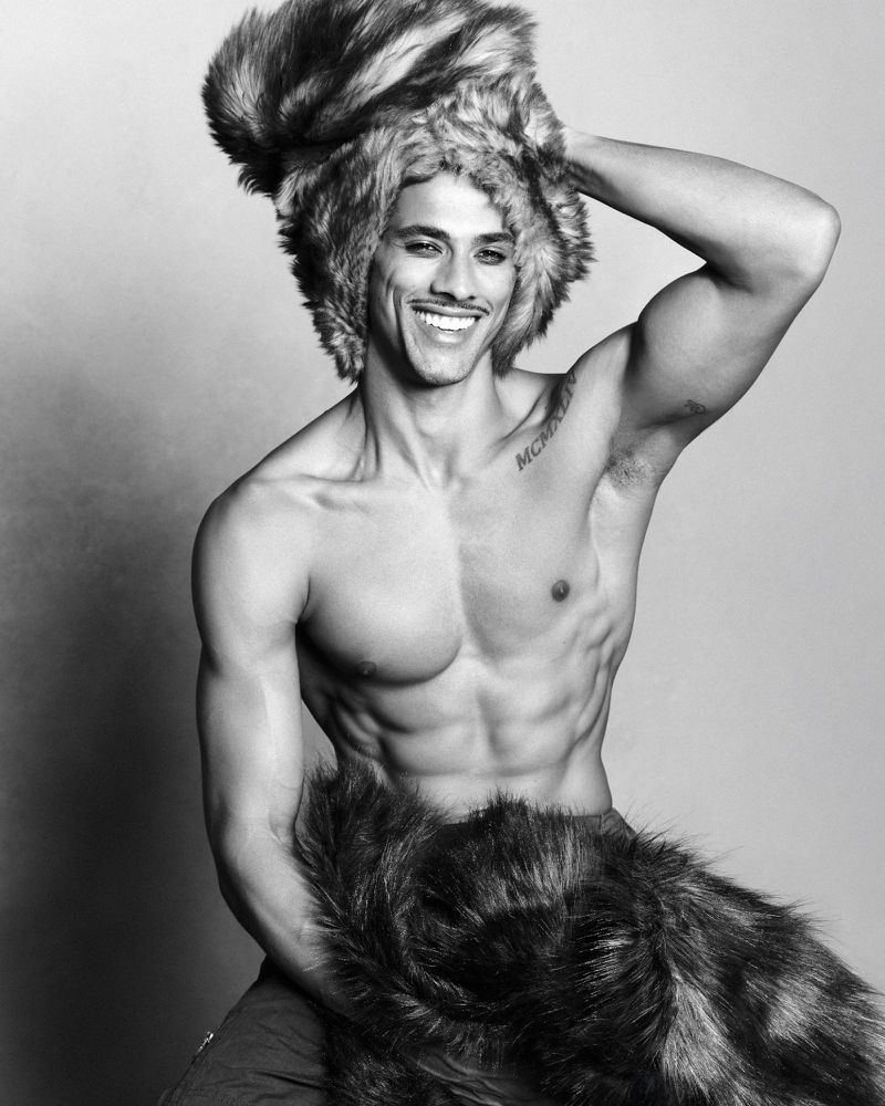 Monochrome portrait of a smiling, muscular man wearing a fur hat and fur wrap against a neutral background in a studio setting.