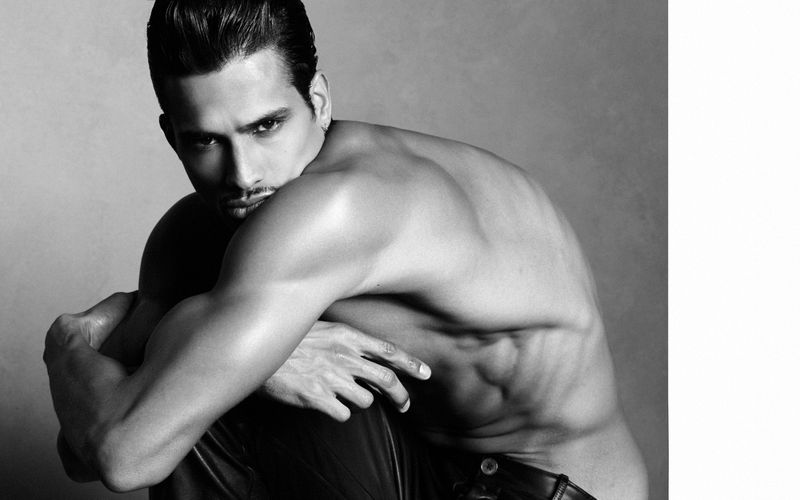 Black and white portrait of model Jonas Barros crouching shirtless against a neutral studio background.