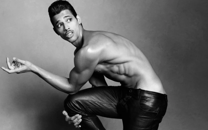 Monochrome studio portrait of model Jonas Barros kneeling shirtless wearing dark trousers against grey backdrop.