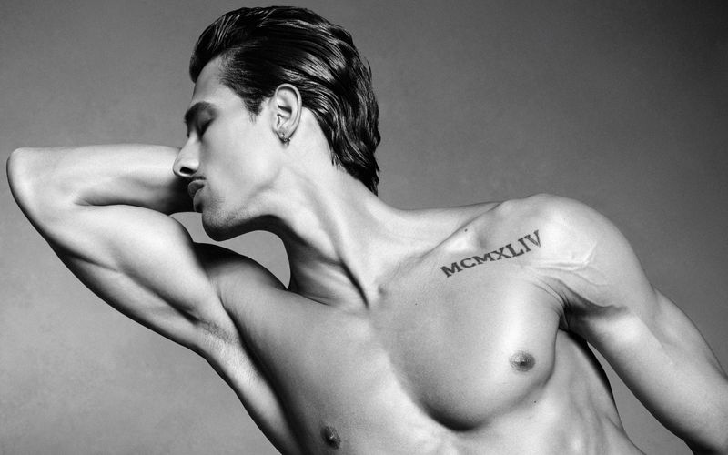 Black and white portrait of male model Jonas Barros flexing arm with Roman numeral tattoo against a plain grey background