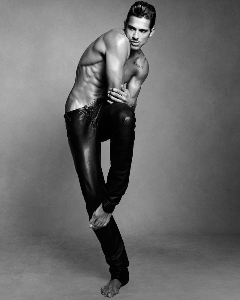 Shirtless male model Jonas Barros wearing dark trousers in a studio portrait against a grey seamless background