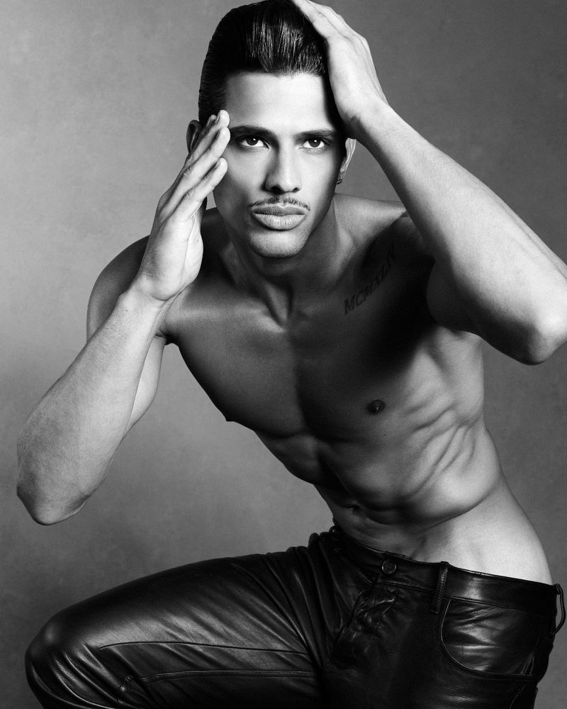 Shirtless male model with dark hair and mustache posing with hands framing face against grey studio background
