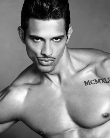 Close-up portrait of shirtless male model Jonas Barros with a mustache and chest tattoo against a plain grey background