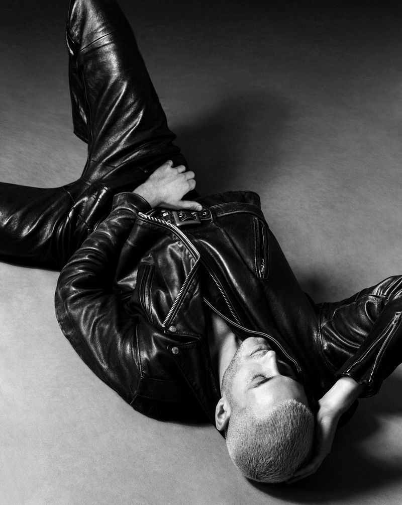 Model Fai Khadra wearing a black leather jacket and leather trousers reclining on a grey studio backdrop.