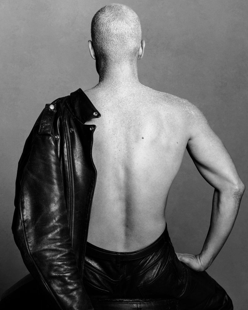 Model Fai Khadra from behind wearing a leather jacket draped over one shoulder against a grey background.