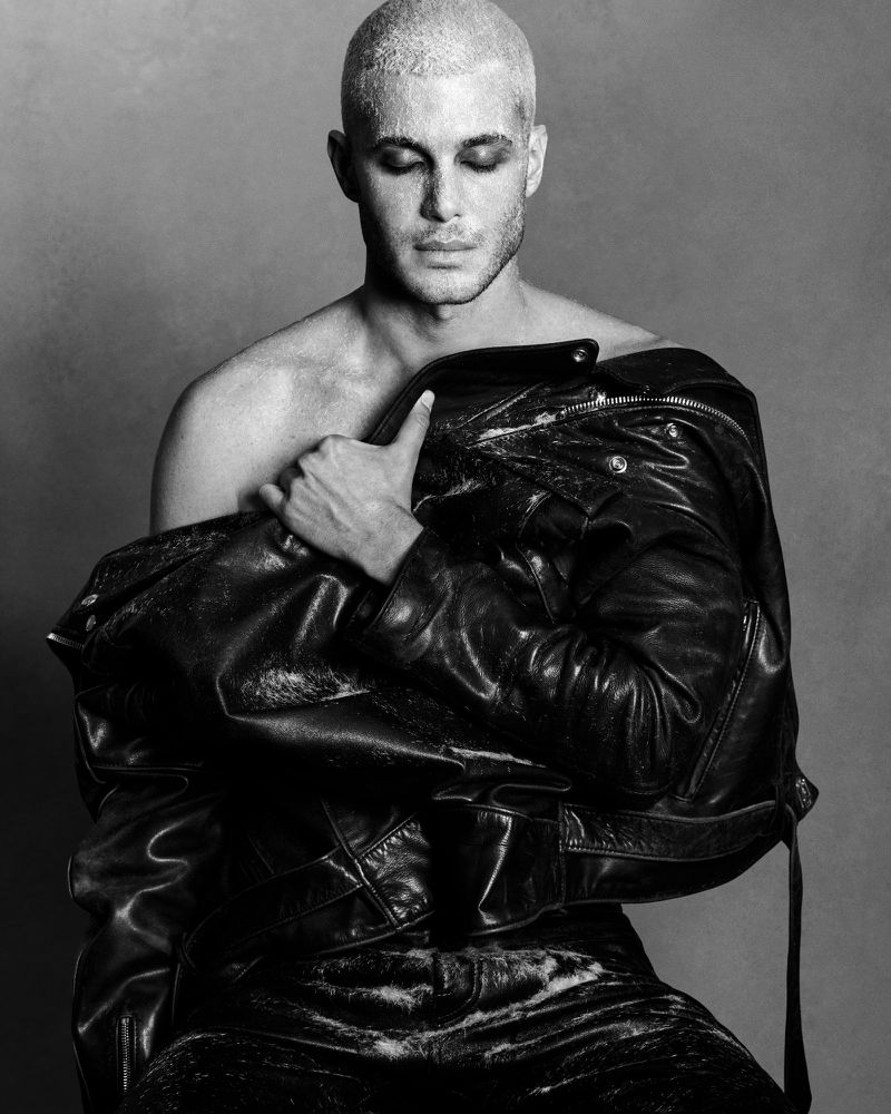Monochrome portrait of Fai Khadra holding a black leather jacket against a plain grey studio background.