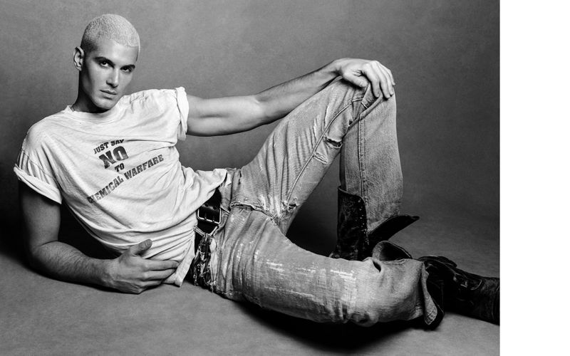 Fai Khadra in a white graphic t-shirt and distressed denim jeans posing against a grey seamless studio backdrop.