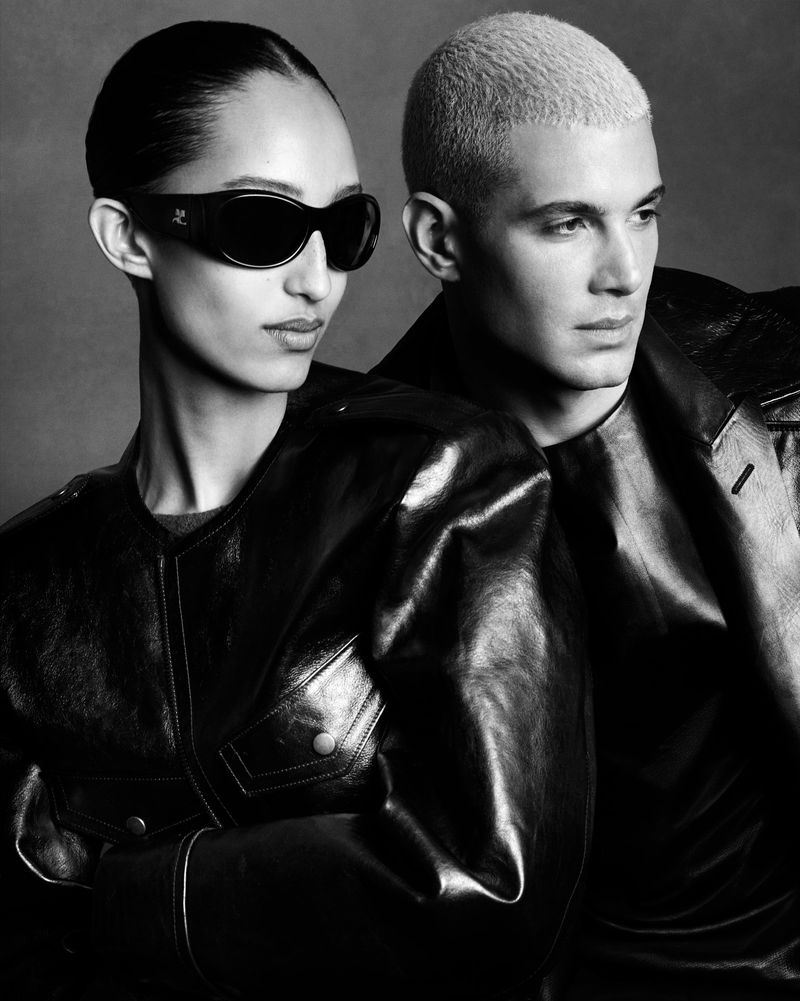 Monochrome fashion portrait featuring two models wearing dark leather jackets against a textured grey background.