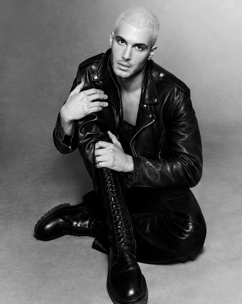 Monochrome portrait of Fai Khadra seated on a seamless backdrop wearing a black leather jacket and tall lace-up boots.