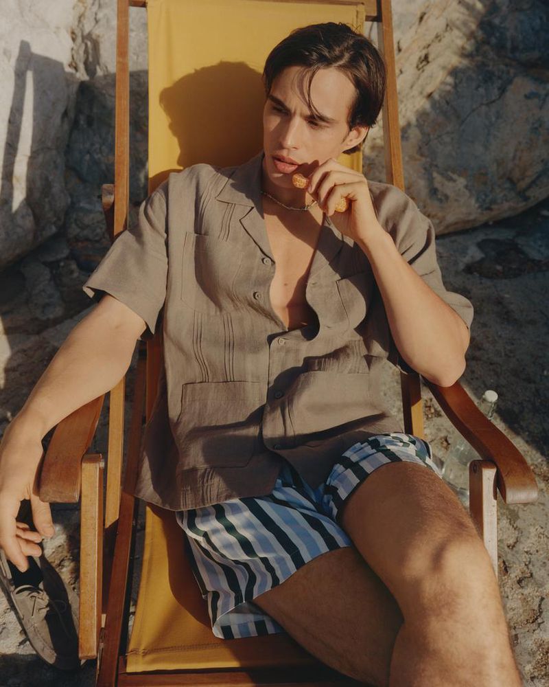A young man seated in a yellow deck chair wearing a grey camp collar shirt and blue striped swim shorts