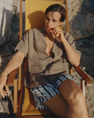 A young man seated in a yellow deck chair wearing a grey camp collar shirt and blue striped swim shorts