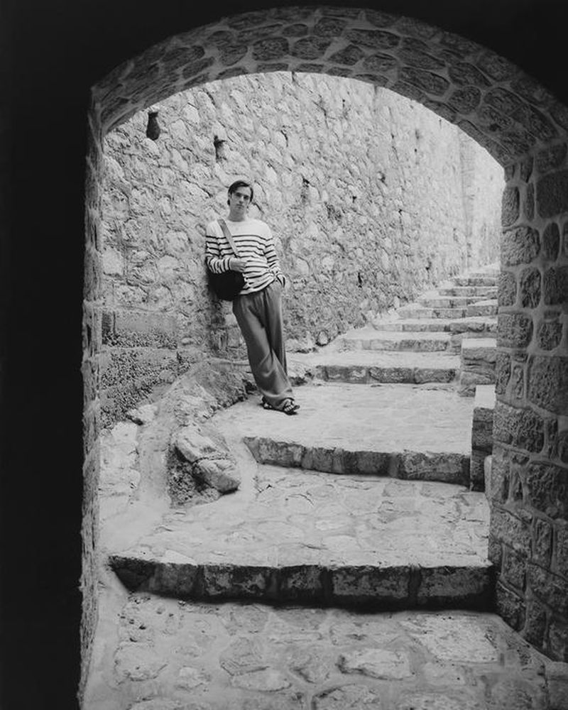 Man in striped shirt leans against stone wall under archway with steps leading down. Black and white photo in outdoor setting.