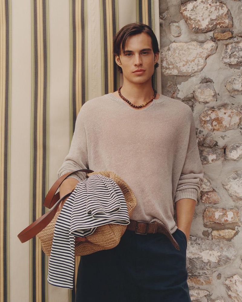 Male model Louis Baines wearing a beige knit sweater and dark trousers holding a straw bag for Zara Man Spring 2026