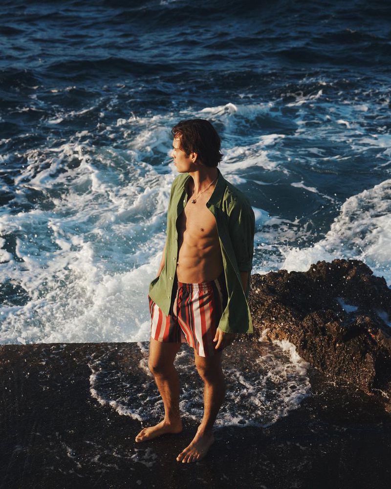 Model Louis Baines standing on rocky shore wearing open olive green shirt and striped shorts by the sea for Zara