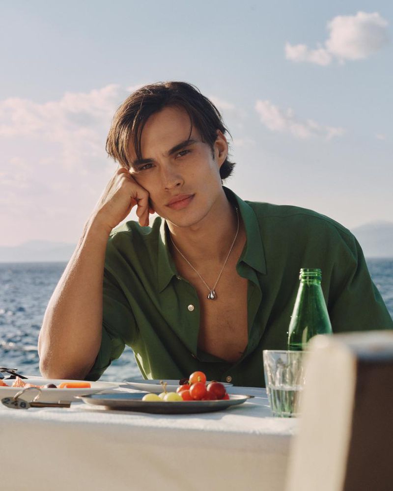 Model Louis Baines seated outdoors at a seaside table wearing a green shirt for Zara, photographed by Ronan Gallagher.