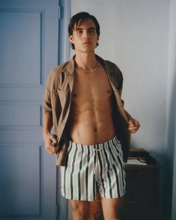 Attractive brunette man in an open brown linen shirt and striped shorts poses indoors against a blue door and white wall.
