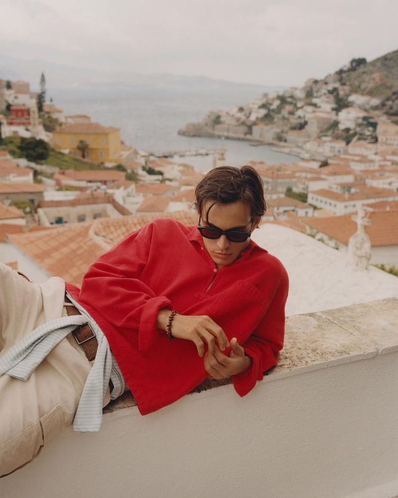 Model wearing a red shirt and sunglasses leaning on a stone wall overlooking a coastal town with tiled roofs.